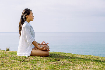 Mindful Asian woman meditating in Lotus pose on green shore