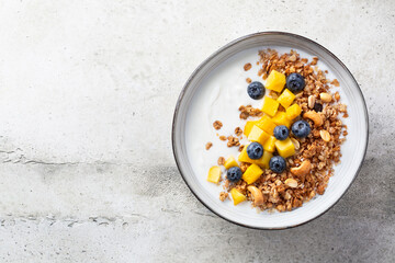 Fototapeta na wymiar Granola (muesli) and yogurt in bowl, nuts, fresh berries blueberry and mango fruit. Healthy eating. Easy breakfast or snack. Top view, copy space, light grey background.