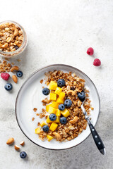 Granola (muesli) and yogurt in bowl, nuts, fresh berries blueberry and mango fruit. Healthy eating. Easy breakfast or snack. Top view, copy space, light grey background.