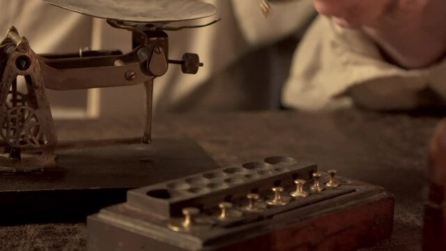 A 19th century Apothecary selecting a weight for the balance scale he utilizes to measure ingredients used in his remedies.