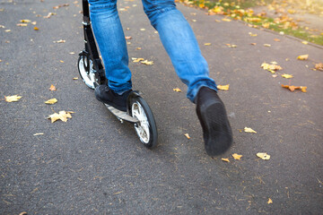 The legs of a man in jeans and sneakers on a scooter in the park in autumn with fallen dry yellow leaves on the asphalt. Autumn walks, active lifestyle, eco-friendly transport, traffic