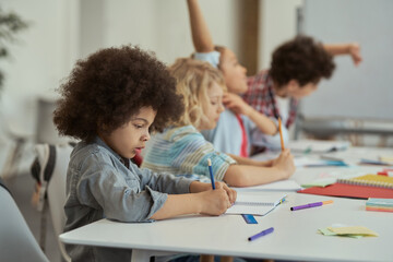 Diligent little schoolboy writing in his notebook while studying, sitting at the table in elementary school classroom