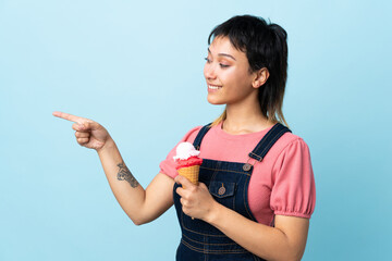 Young Uruguayan girl holding a cornet ice cream over isolated blue background pointing to the side...