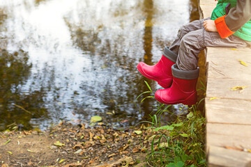 A child in red rubber boots sits on a wooden bridge over the river and looks at the water. Autumn landscape, cold weather, dry fallen leaves. Autumn mood, carefree childhood