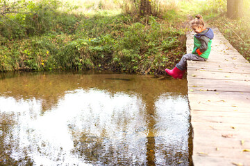 Fototapeta na wymiar A child in red rubber boots sits on a wooden bridge over the river and looks at the water. Autumn landscape, cold weather, dry fallen leaves. Autumn mood, carefree childhood