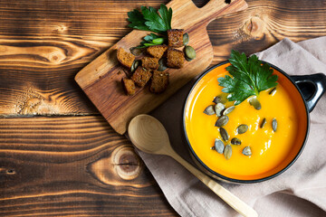 Pumpkin soup in a bowl with seeds and parsley on a wooden background. Crackers with spices on the board. Copy space. Thanksgiving, autumn soup
