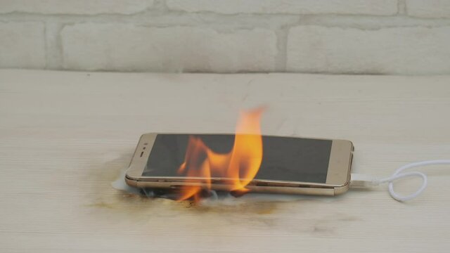 cell phone with swollen battery burns with red flame while charging while lying on desk at home or in office. Concept of cause of fire in house.