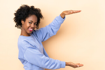 Young African American woman isolated on beige background holding copyspace to insert an ad