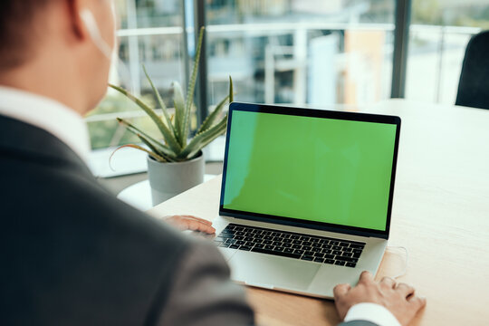 businessman in suit in front of laptop with green screen has his hands on his near keyboard while being at video conference or surfing online at web with copy space and mockup possibility on monitor