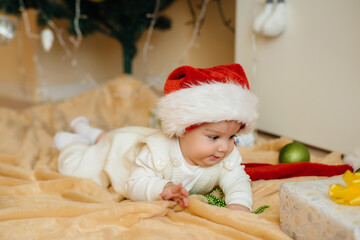 Cute smiling baby is lying under a festive Christmas tree and playing with gifts. Christmas and New Year celebrations