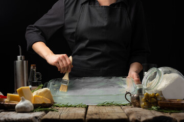 The cook greases a baking dish with olive oil. Preparation of traditional Italian focaccia bread. Close-up, wood texture. Ingredients. Italian cuisine, cookery. bakery, cookbook.