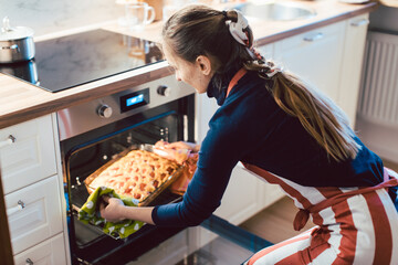 Beautiful young woman wearing apron removing baked apple pie from microwave oven in kitchen at home