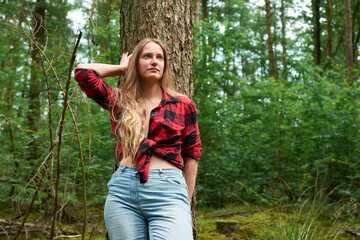 A young Italian woman wearing a red flannel with jeans and leaning on a tree in a forest - obrazy, fototapety, plakaty