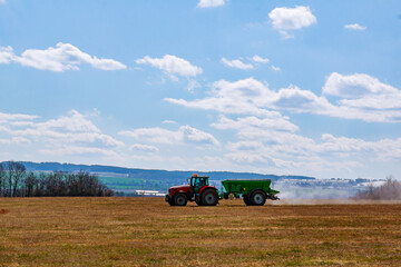 Obraz premium A red tractor with a green trailer spreads granular fertilizer on the ground in the field.