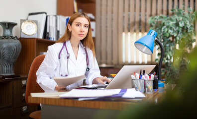 Female doctor with stethoscope on shoulders sitting at desk in office and using laptop.