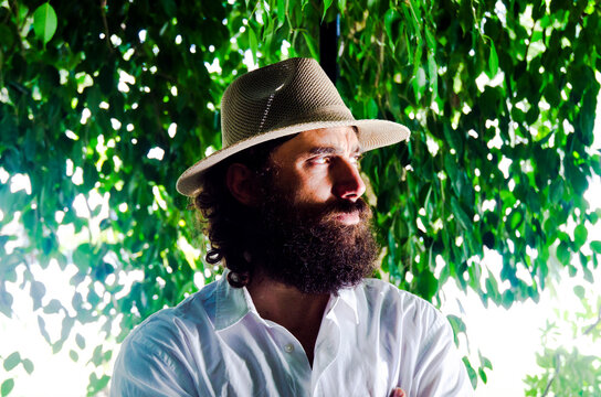 young man with beard and long hair wears hat and white shirt surrounded by greenery