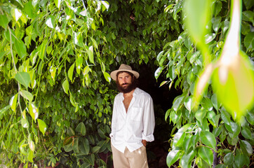 young man with beard and long hair wears hat and white shirt surrounded by greenery