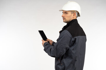 Worker with a smartphone in his hands. Working man with smartphone. Portrait of a worker on a white background. He looks away. Concept worker wants to call someone. Man in a gray uniform and a helmet