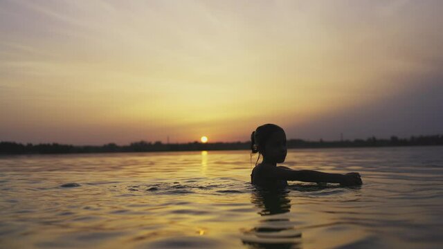 A girl plays with water in the lake and splashes it to the sides against the backdrop of a sunset