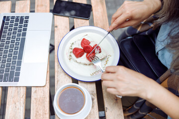 Senior woman eats delicious toast with fresh strawberries and cream near open laptop at table on outdoors cafe terrace closeup