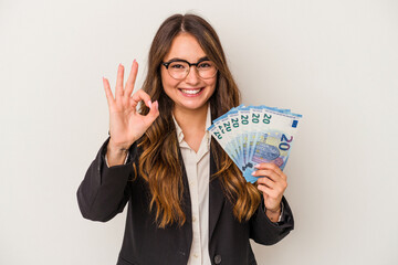 Young caucasian business woman holding banknotes isolated on white background cheerful and confident showing ok gesture.