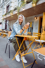 Thoughtful senior Asian woman with silver hair and mobile phone sits at small yellow table on outdoors cafe terrace on autumn day
