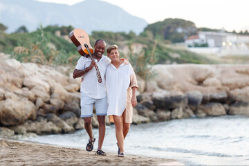 Happy mixed race, middle age couple embracing while walking on the beach holding a guitar. Man...