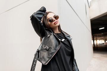 Urban beautiful young hipster woman with stylish round sunglasses in a fashion leather jacket with...
