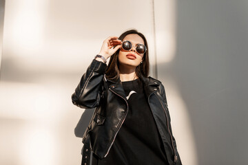 Trendy hipster young woman in fashion black clothes with stylish leather jacket looks through...