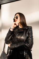 Obraz premium Beautiful stylish hipster girl with vintage sunglasses in a fashion leather jacket with a fashionable black handbag walks in the city at sunset