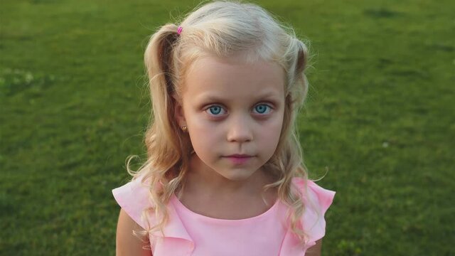Little girl in a pink dress close up. She stands on the green grass in the park and smiles sincerely. She has blonde wavy hair and honest blue eyes