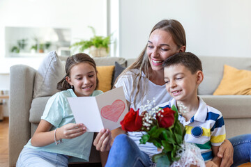 Young mother with a bouquet of roses laughs, hugging her son, and сheerful girl with a card and roses congratulates mom during holiday celebration in kitchen at home