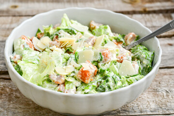 Homemade traditional Caesar Salad with Parmesan Cheese and Croutons in a white bowl on a old white table with leaves and a sauce boat 