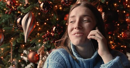 Close up head shot happy 30s young woman holding mobile phone conversation, congratulating friends or family with New Year or Merry Christmas winter holidays, saying best wishes near decorate tree.