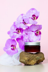 A brown jar of cream on a pink background stands on a large building stone with beautiful orchids next to it. Stylish appearance of the product, layout, personality.