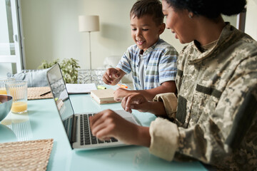 Boy looking at the laptop while his soldier mother in military uniform working