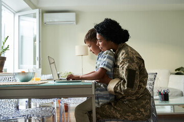 Woman embracing her little son sitting at her laps and typing at the laptop keyboard