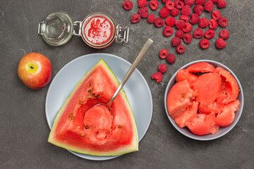 Spoon in pulp of ripe watermelon. Pieces of watermelon in gray bowl. Watermelon juice in glass jar. Apple and raspberry on table