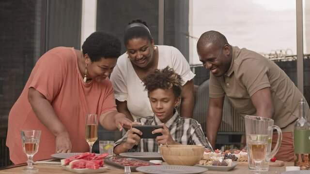 Medium long of teenage African boy using smartphone, sitting at dinner table, his parents and grandmother standing behind him, looking at him, talking and smiling