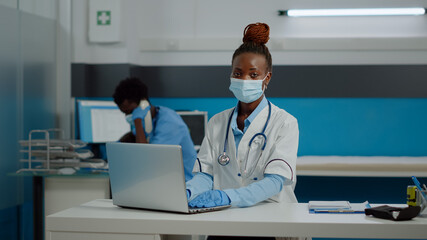 Portrait of young medic using laptop technology on desk in healthcare office at clinic. Doctor working with modern gadget in medical cabinet wearing white coat, gloves and face mask