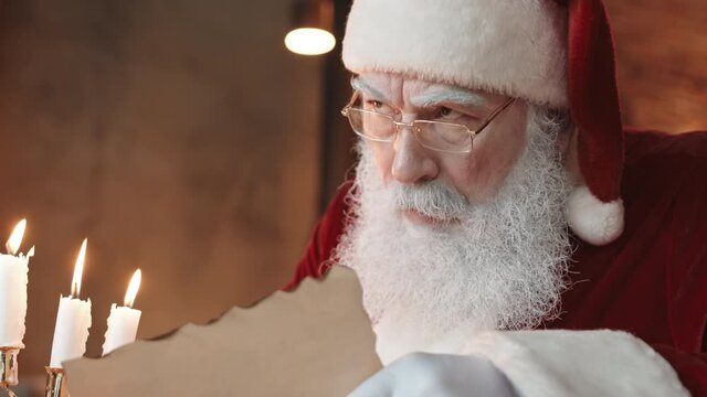 Medium close-up of serious Santa Claus wearing red and white costume, sitting at home, reading letter, nodding and thinking