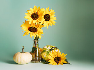 Yellow Gerbera flowers in a glass vase surrounded by autumn gourds and pumpkins on pastel green background. Creative fall concept. Autumn natural still life wallpaper. © Aleksandar