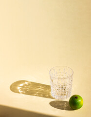 an art creative template of empty crystal glass and lime under hard sunlight, glitter reflection on beige background, long harsh rectangular and oval shadows, copy space