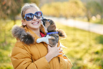 Girl in sunglasses holding puppy and smiling in autumn in park. Concept of communication child with pet.
