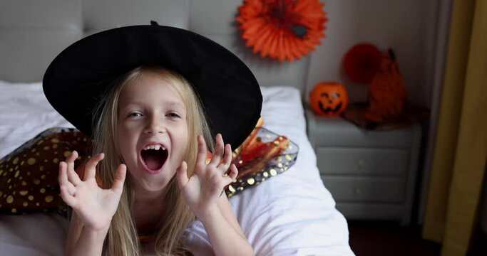 Cute Caucasian little girl with blonde hair seven years old in witch dress and hat having fun and celebrating Halloween at home during Coronavirus covid-19 pandemic and quarantine. Slow motion