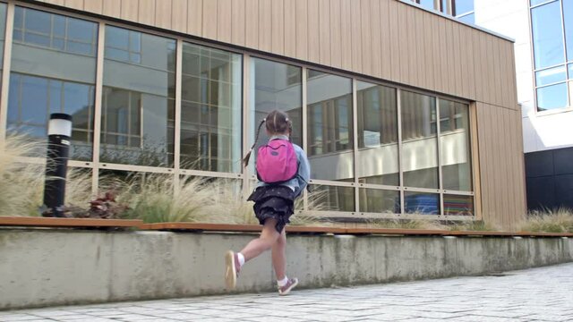 An elementary school student hurries to school. Little girl with pigtails and a knapsack runs against the background of the school building.