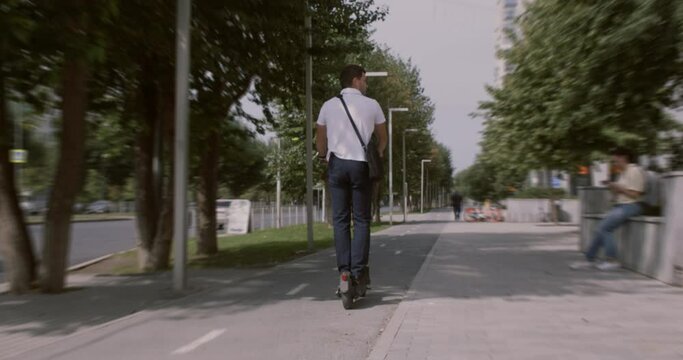 Back view of businessman riding electric scooter in city. Rear view of young man riding electric scooter along pathway on city street