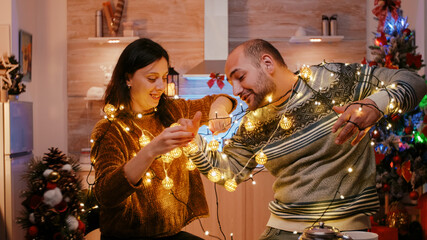 Cheerful couple getting tangled in string of twinkle lights for christmas eve preparations. Man and...