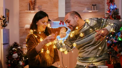 Fotobehang Cheerful couple getting tangled in string of twinkle lights for christmas eve preparations. Man and woman laughing while untangling knot of garland with lights and illuminated bulbs © DC Studio