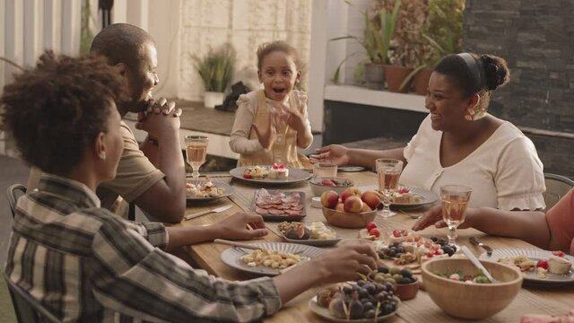 Medium of cheerful African-American family sitting at table in backyard, having conversation, then raising and clinking glasses
